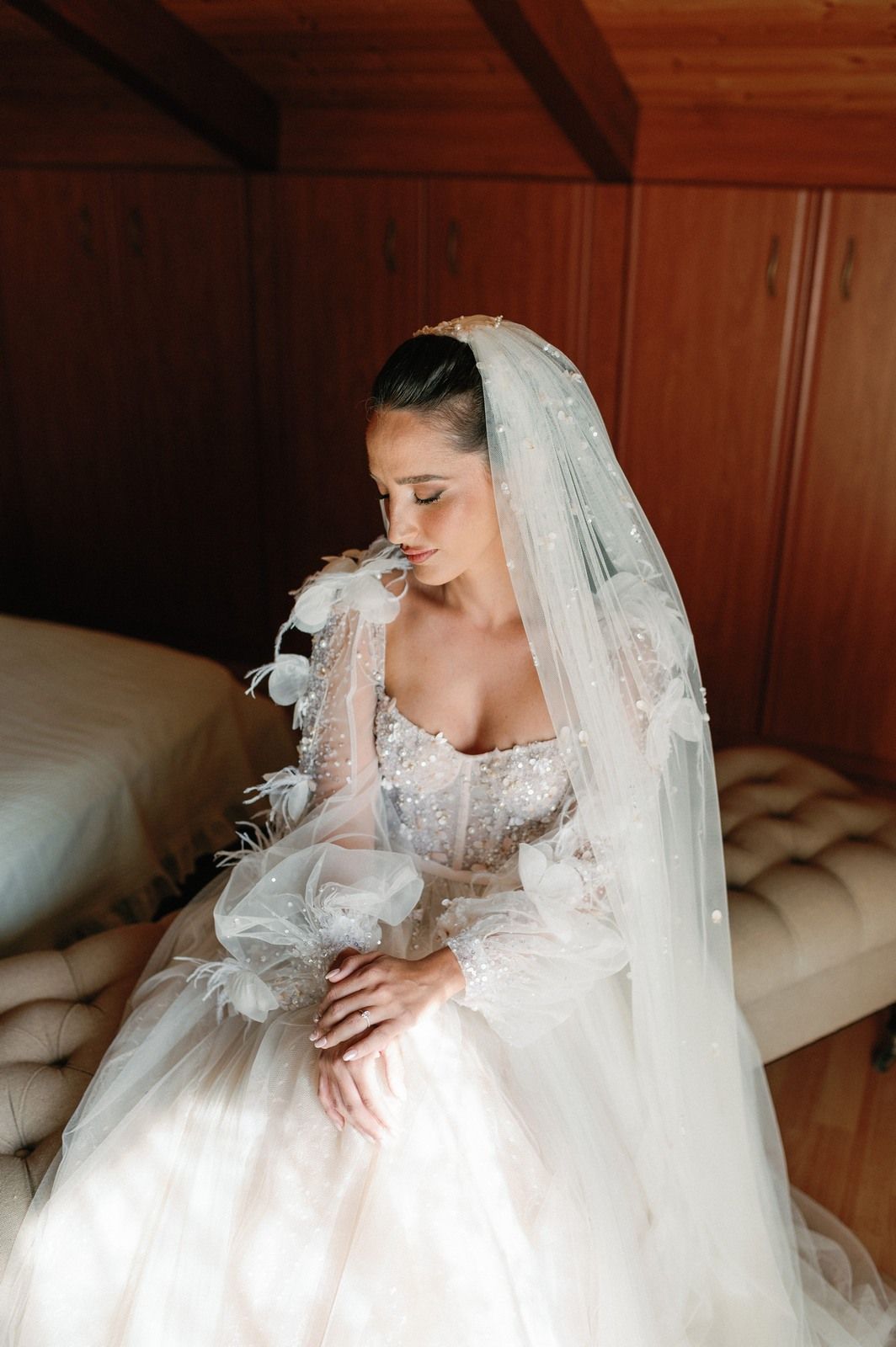 A bride in an elegant white lace wedding gown and veil poses gracefully in a sunlit room.
