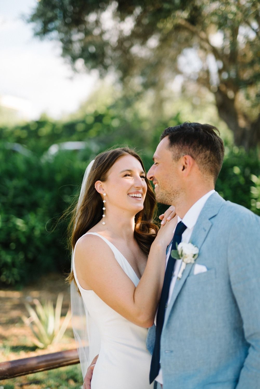A couple in wedding attire share an intimate moment outdoors with natural lighting and greenery in the background.