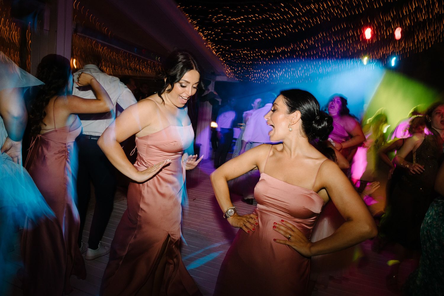 Bridesmaids in salmon-colored dresses strike poses while dancing at a wedding reception.