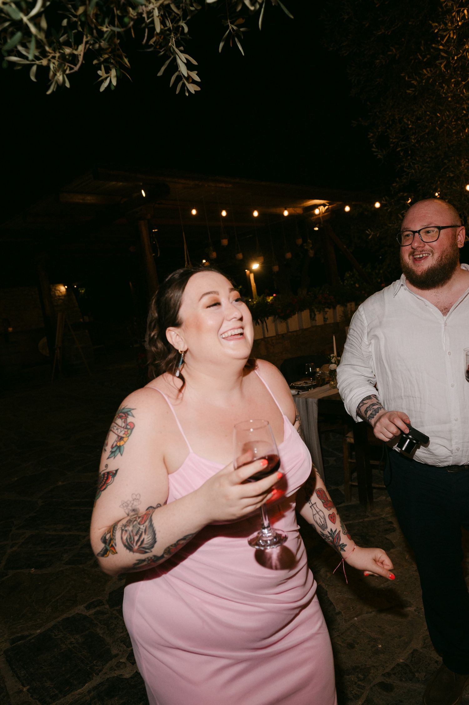 Two wedding guests laugh and enjoy drinks at an outdoor nighttime reception under string lights.