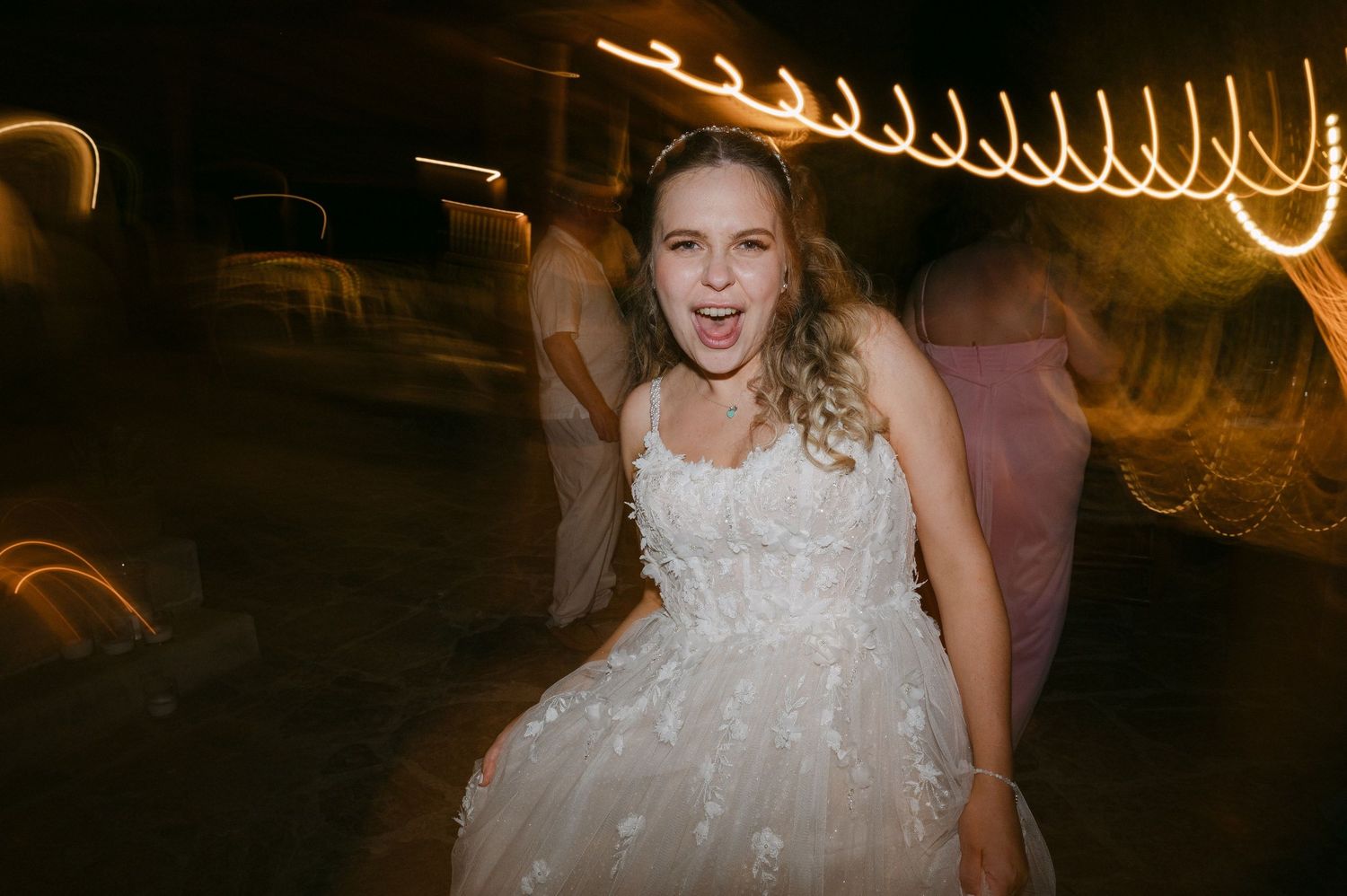 A bride in a white floral wedding dress laughs joyfully at night with blurred light trails swirling behind her.