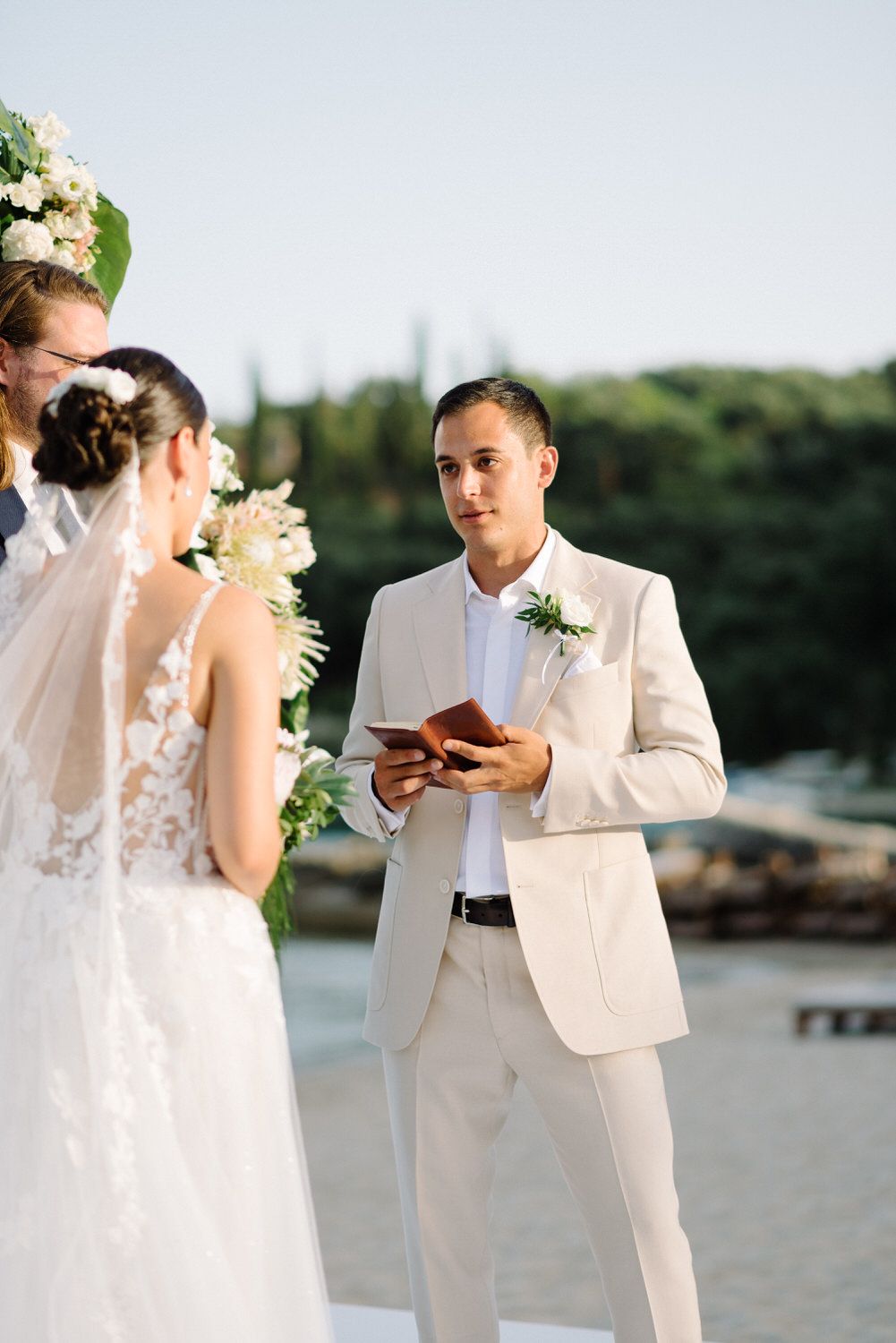 Couple exchanging vows during seaside ceremony with officiant in cream suit.