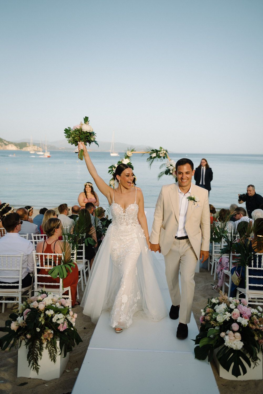 Joyful newlyweds walk down aisle at oceanfront ceremony with guests watching.