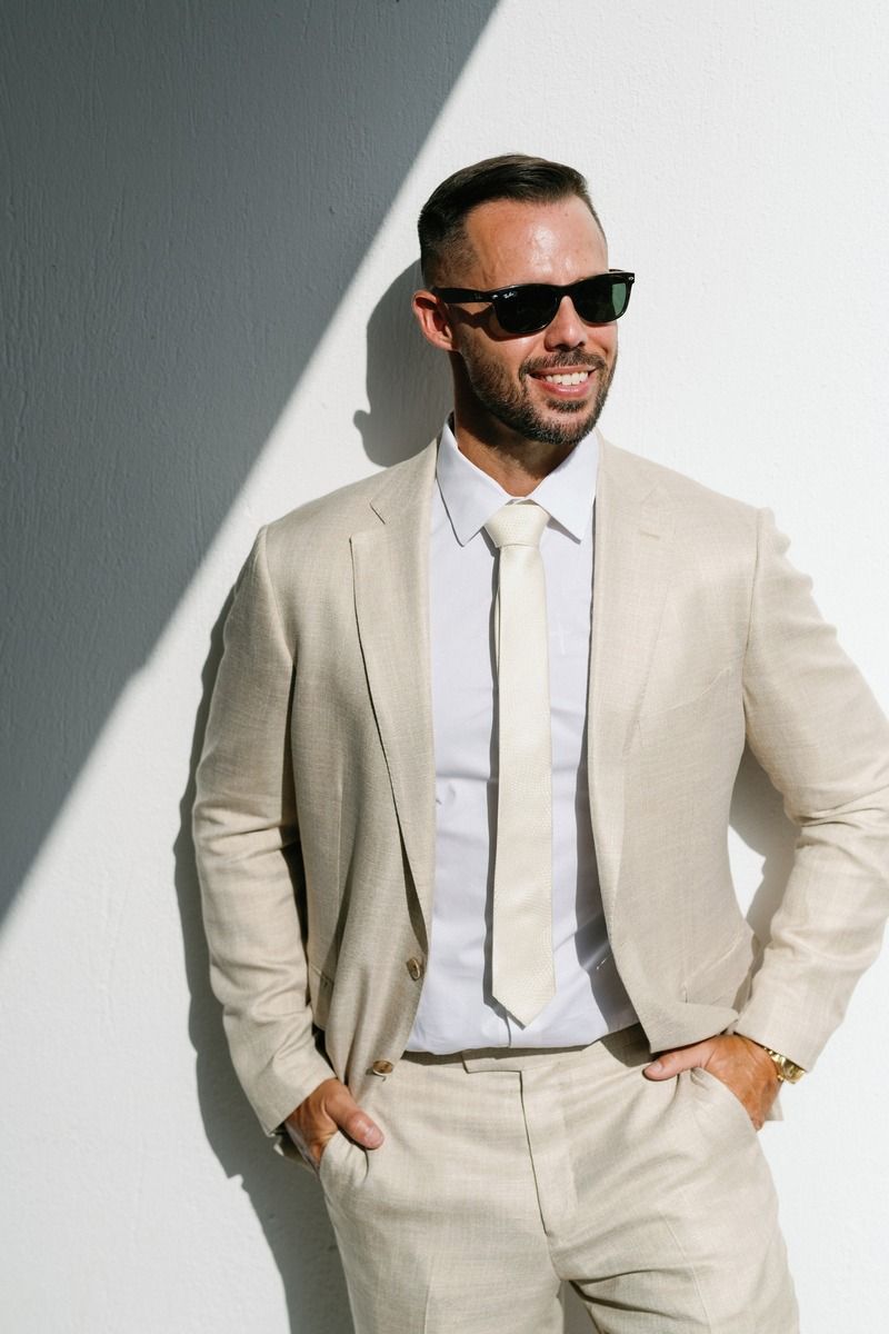 Stylish groom in beige suit and sunglasses poses against white wall with dramatic lighting.