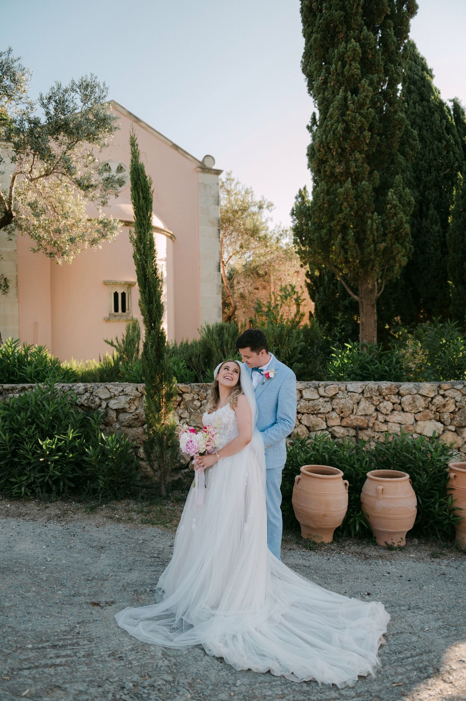 A romantic wedding couple poses together in a Mediterranean garden setting with stone walls and cypress trees.