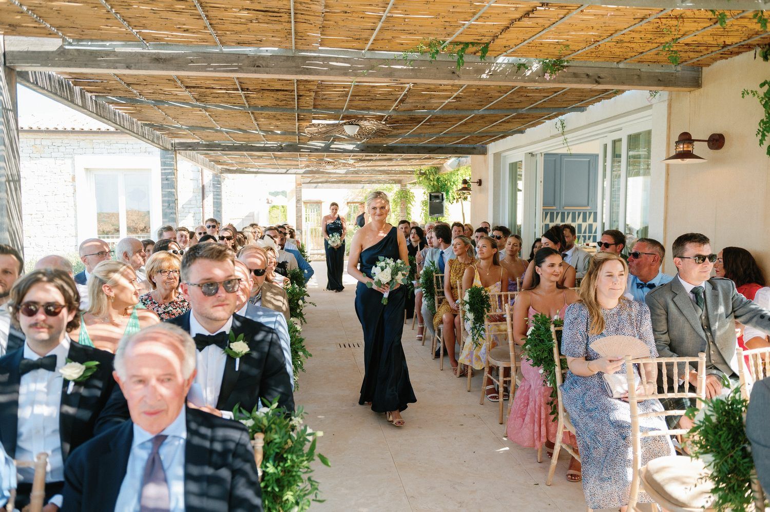 Wedding ceremony processional with guests seated in aisle under bamboo-covered walkway.