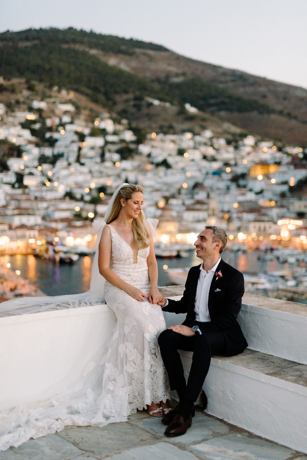 Wedding couple shares romantic moment overlooking Mediterranean coastal town with twinkling lights at dusk.