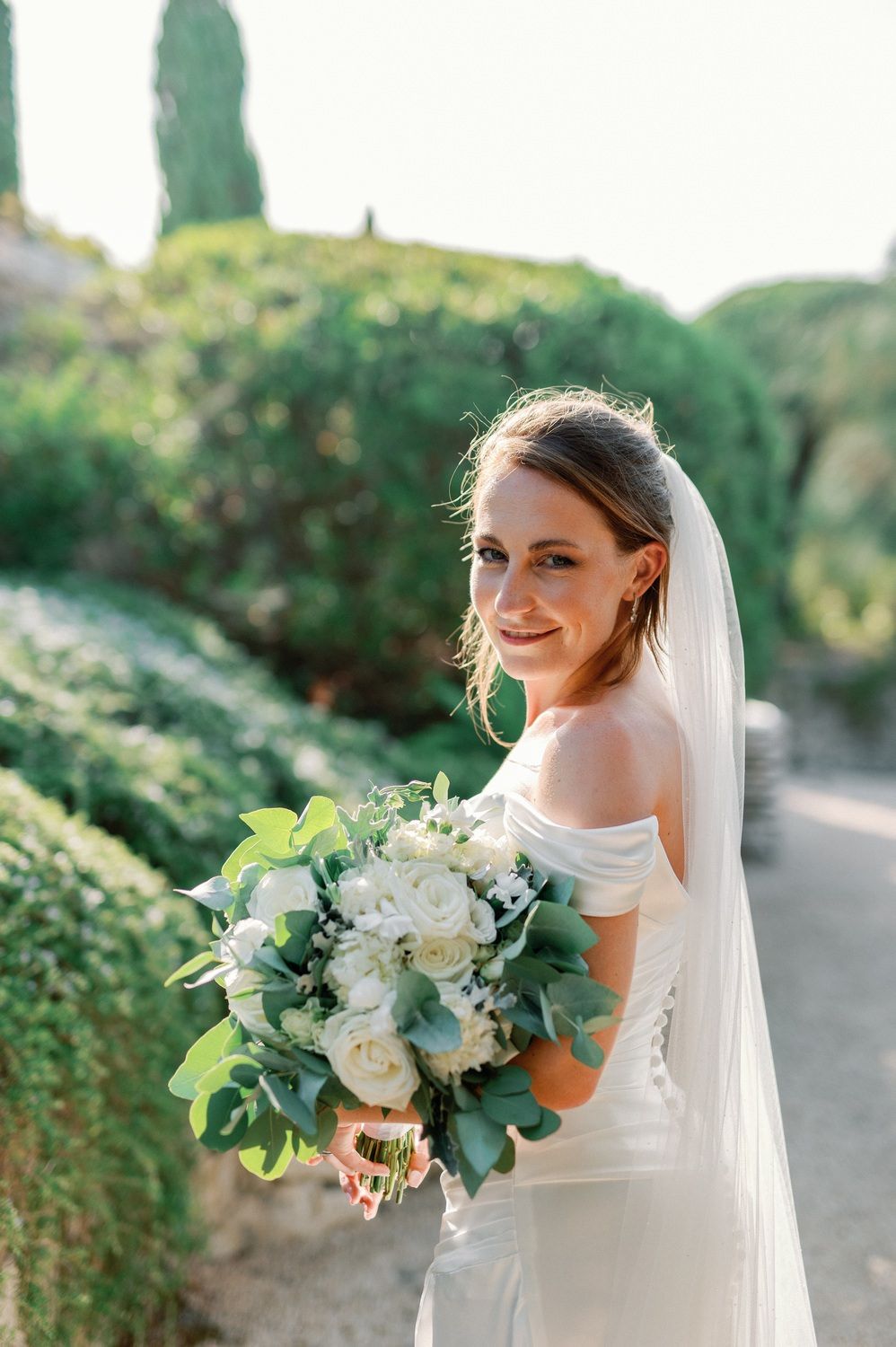 Elegant bride in off-shoulder dress holds lush white and green bouquet in garden setting.