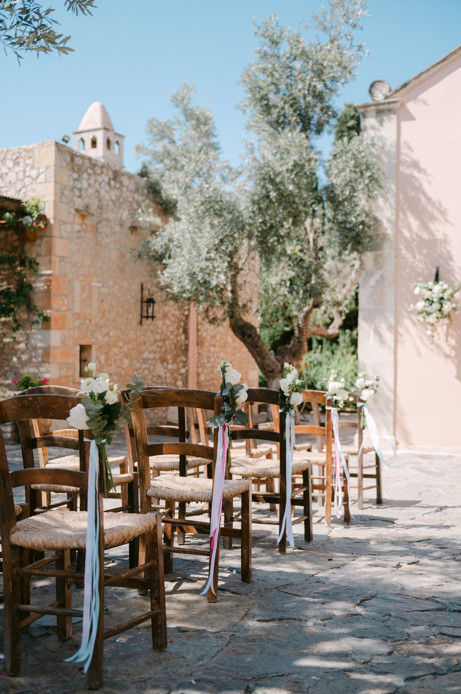 Rustic wooden chairs with soft blue ribbons arranged in rows for an outdoor ceremony at a Mediterranean stone venue.