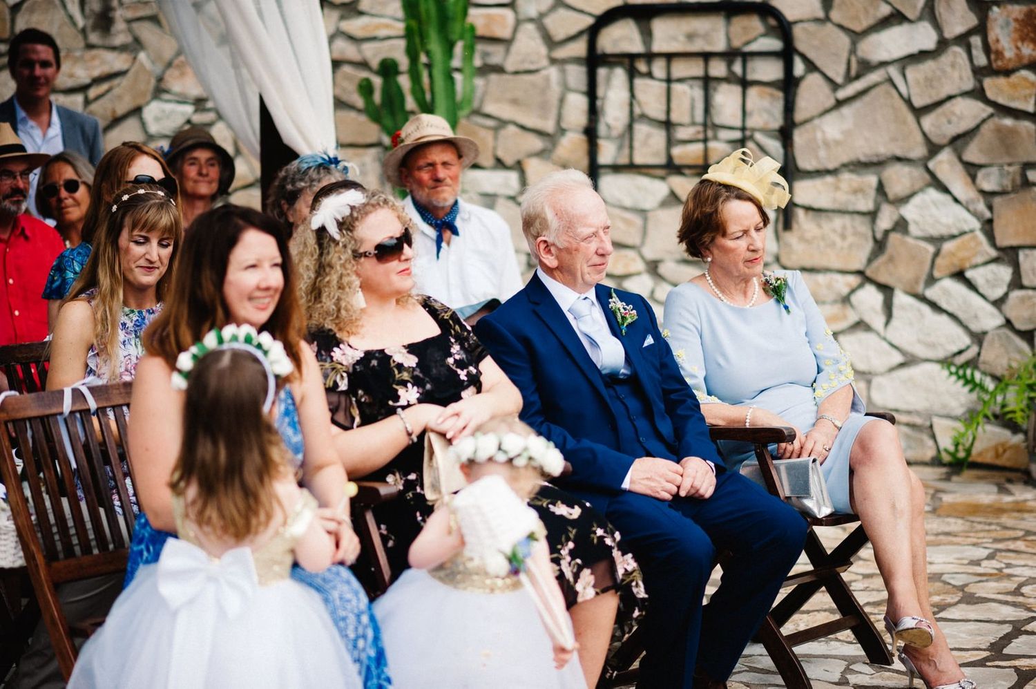Wedding guests seated on wooden chairs watching ceremony at stone patio venue.