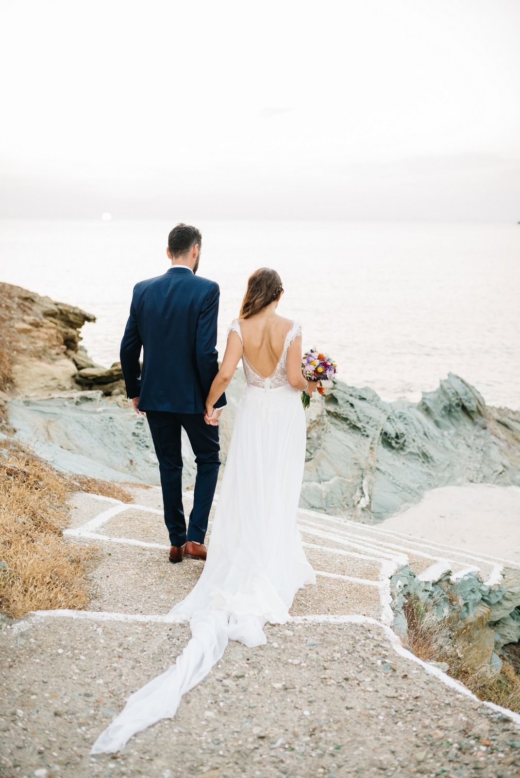 Bride and groom walking along coastal path with flowing white dress trailing behind.