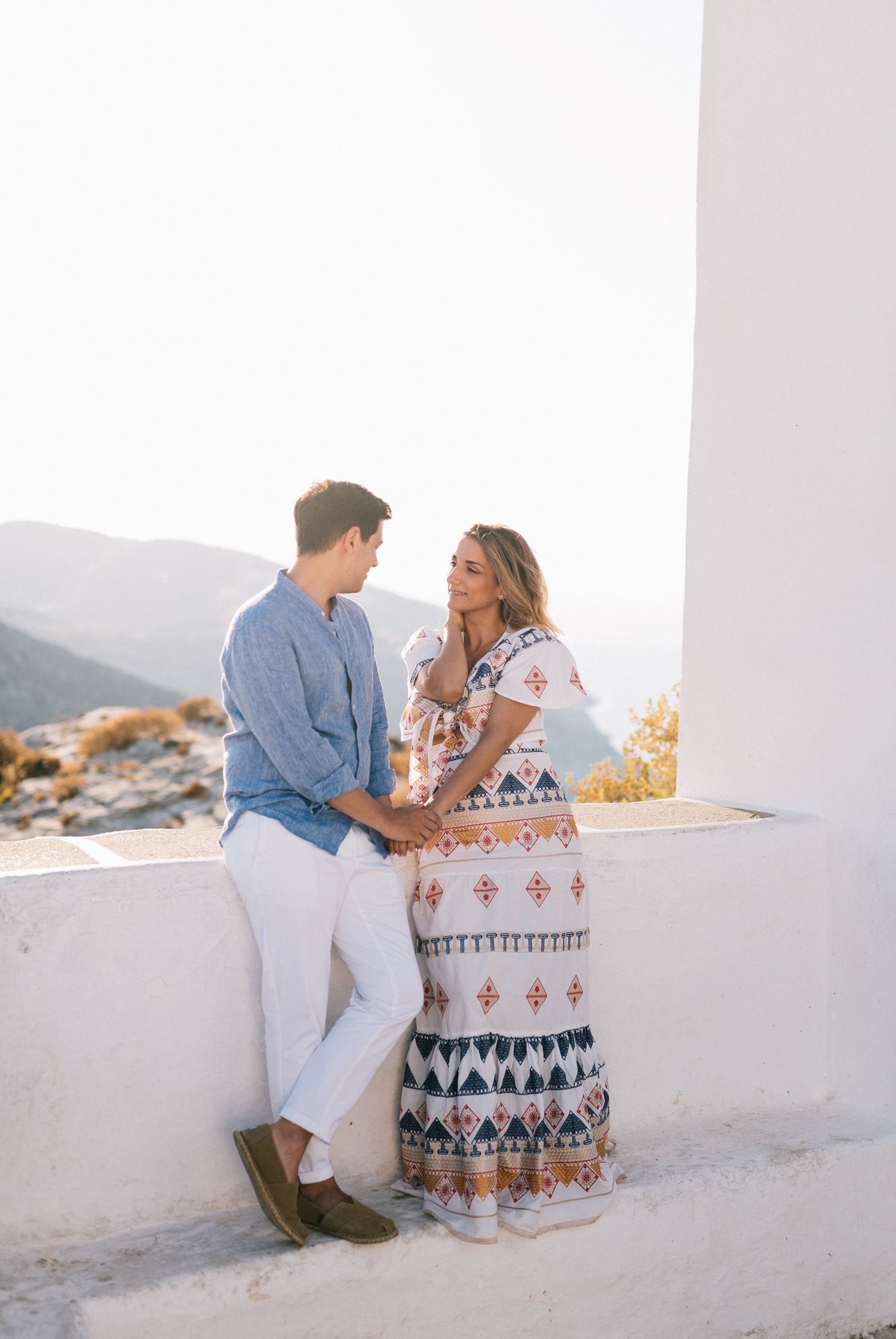 Romantic couple leans on white wall overlooking mountainous landscape at sunset.