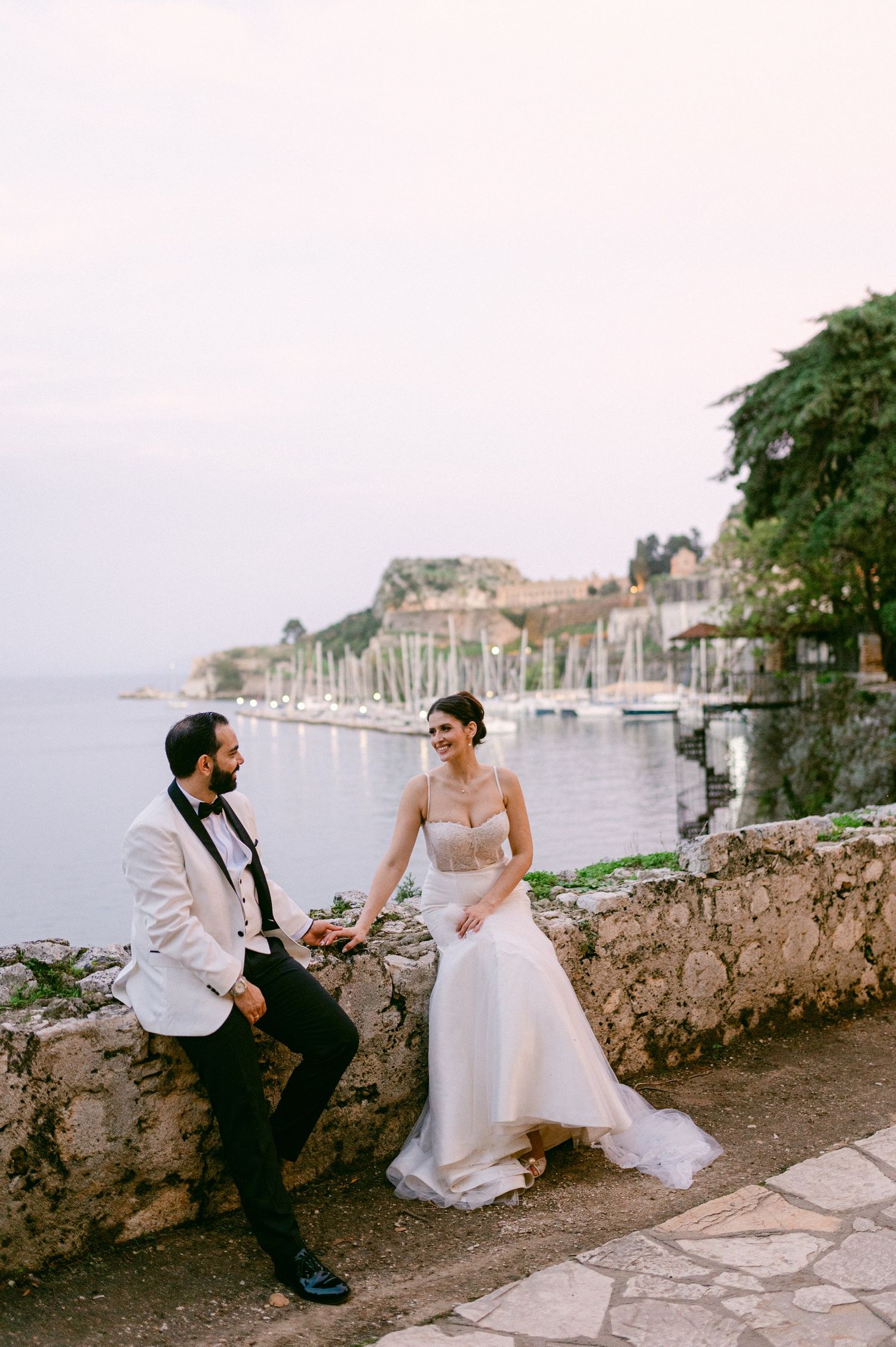 A romantic wedding couple sits on a stone wall overlooking a scenic coastal harbor with boats and buildings in the distance.