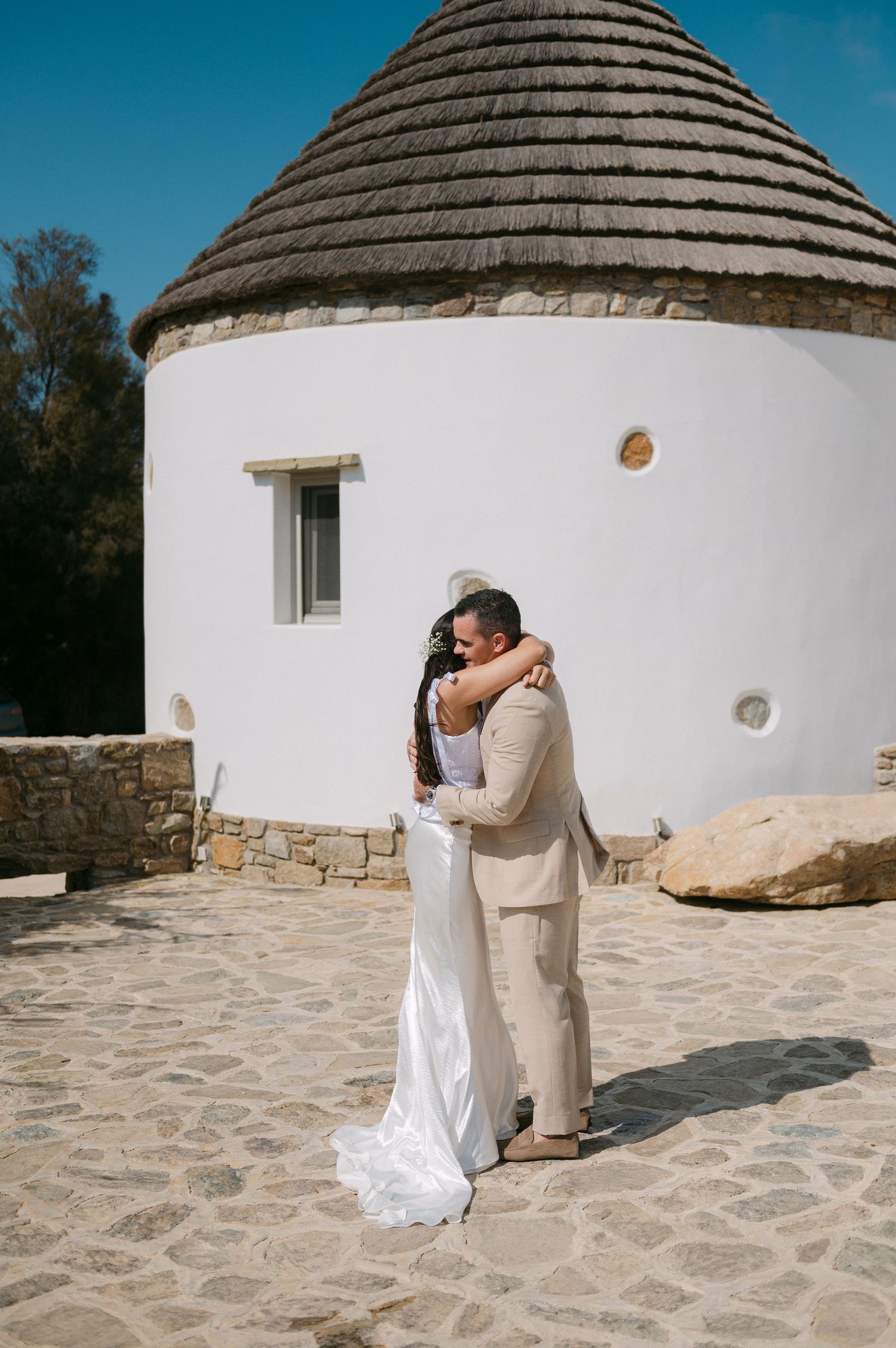 A couple embraces on a stone patio in front of a white circular building with a conical thatched roof.