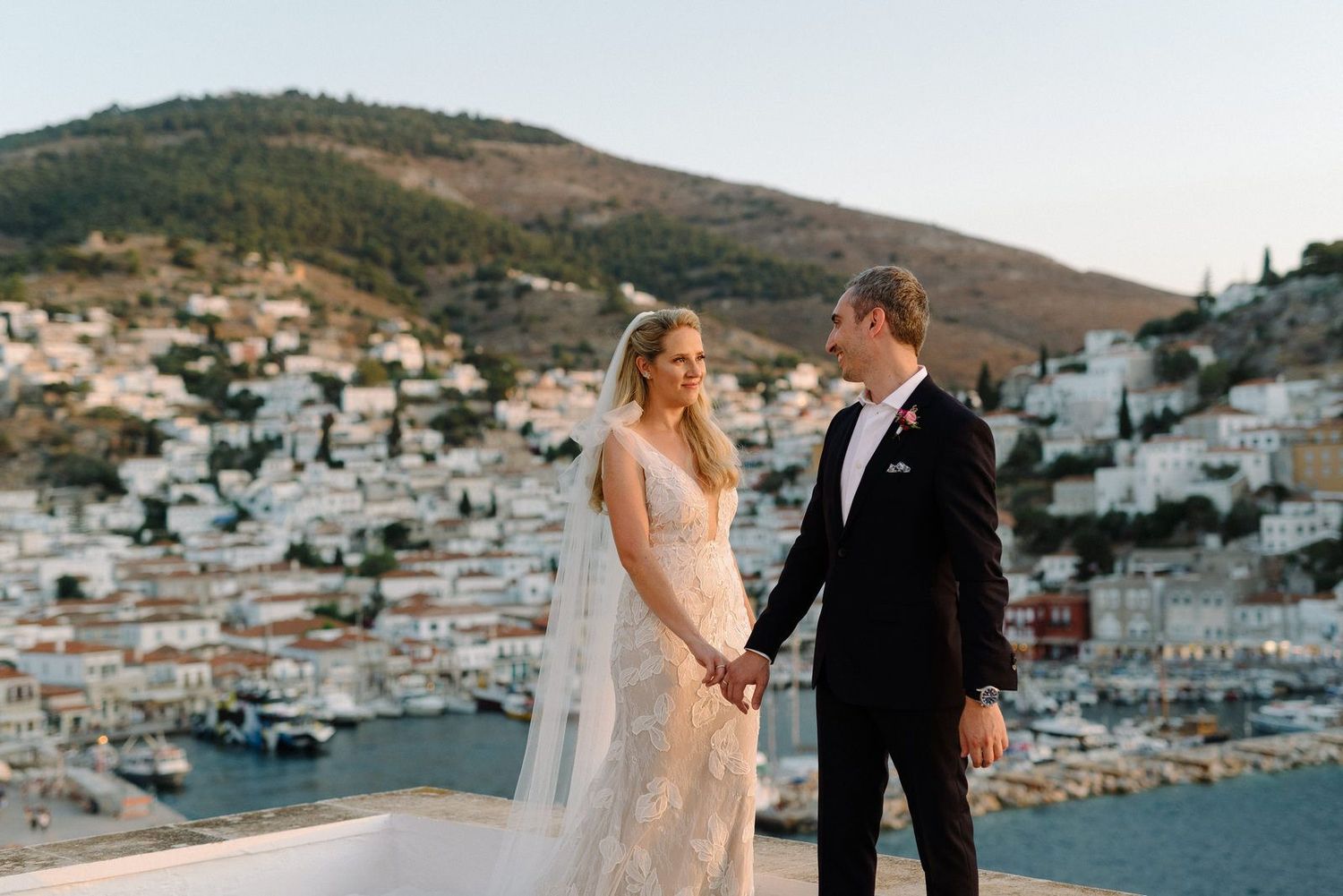 Couple in wedding attire walks along scenic overlook with white hillside village and Mediterranean Sea in background.