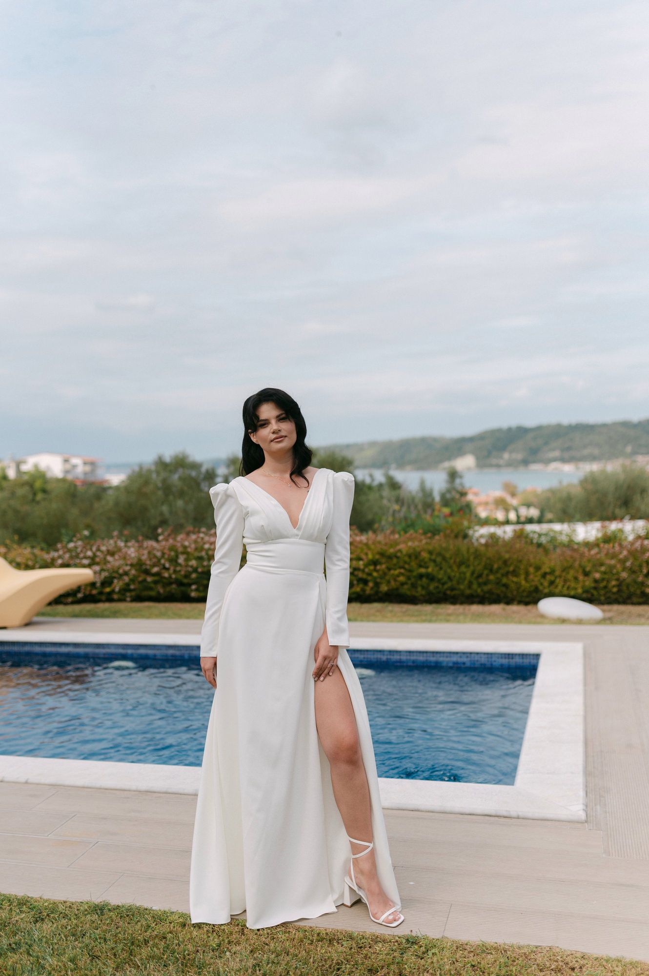 A woman in a flowing white dress poses elegantly by a luxurious poolside with scenic mountain views in the background.