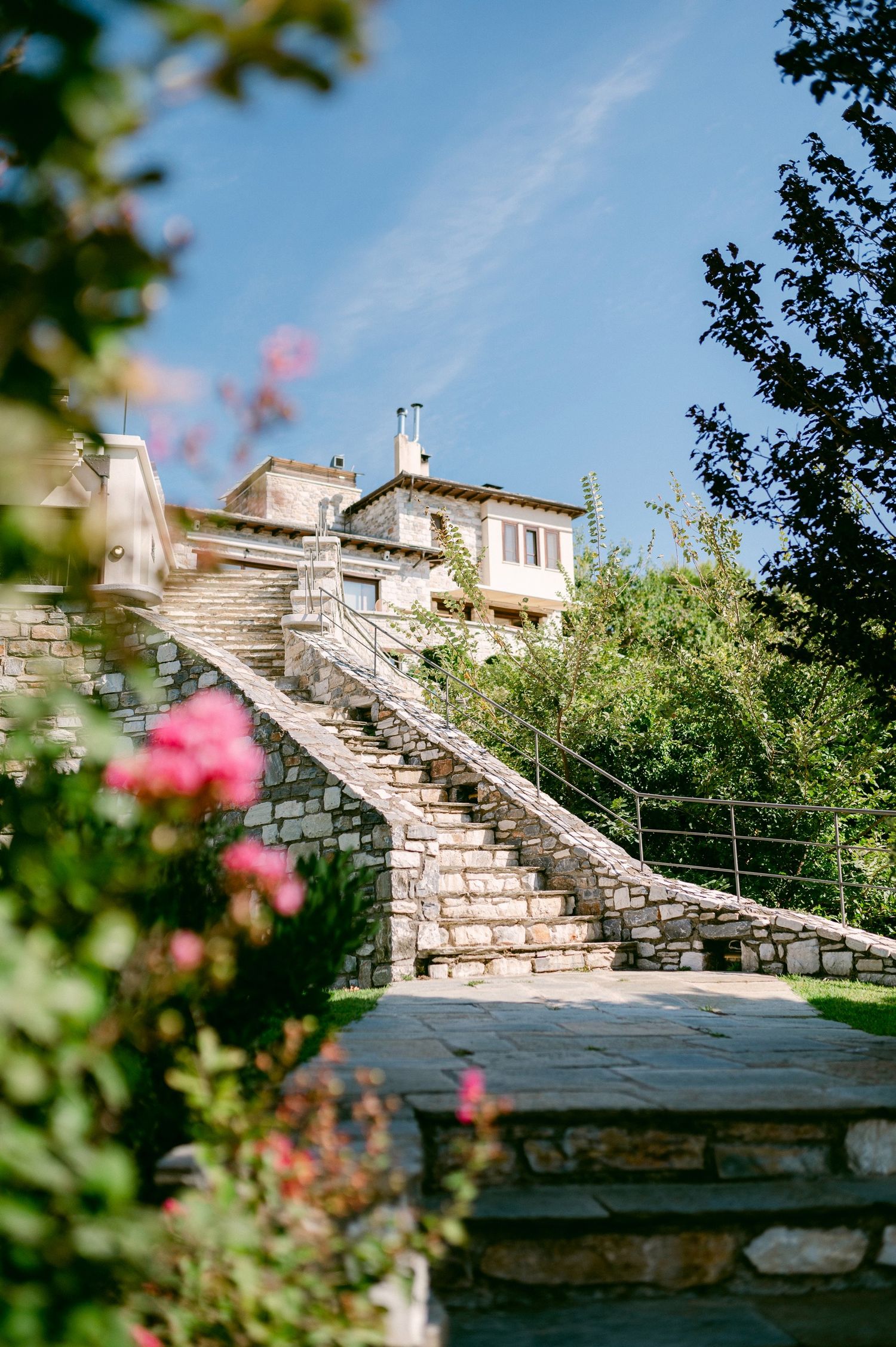 Stone staircase leading to a Mediterranean-style villa with pink flowers blooming in the foreground against a blue sky.