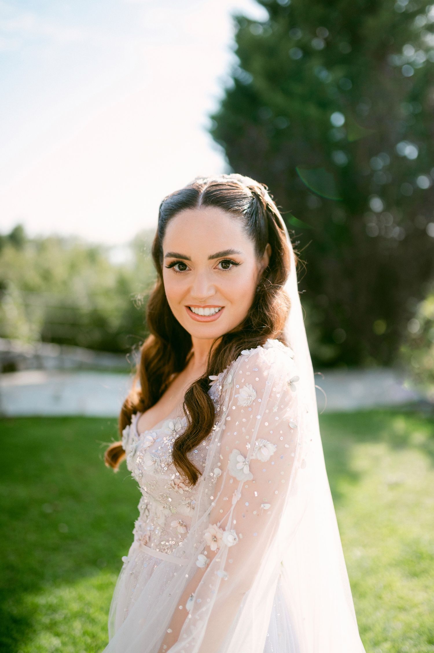 Beautiful bride in lace wedding dress with long veil poses in outdoor garden setting on sunny day.