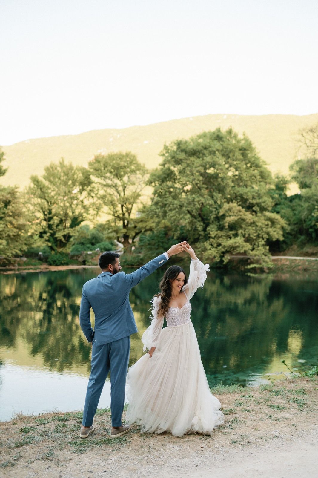 A couple in wedding attire dances together by a serene lake surrounded by trees at sunset.