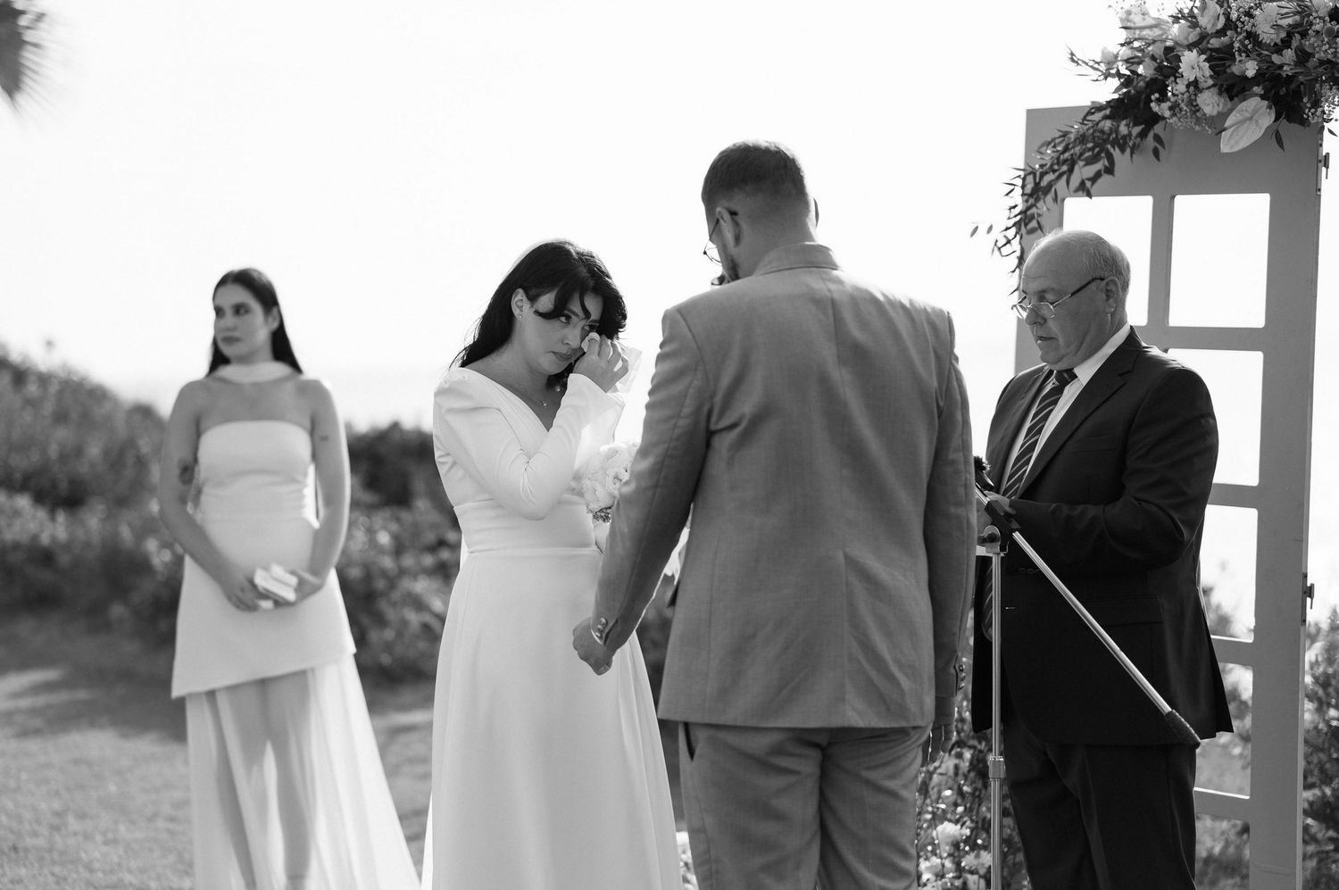 A black and white outdoor wedding ceremony with a couple exchanging vows under an arch with an officiant.