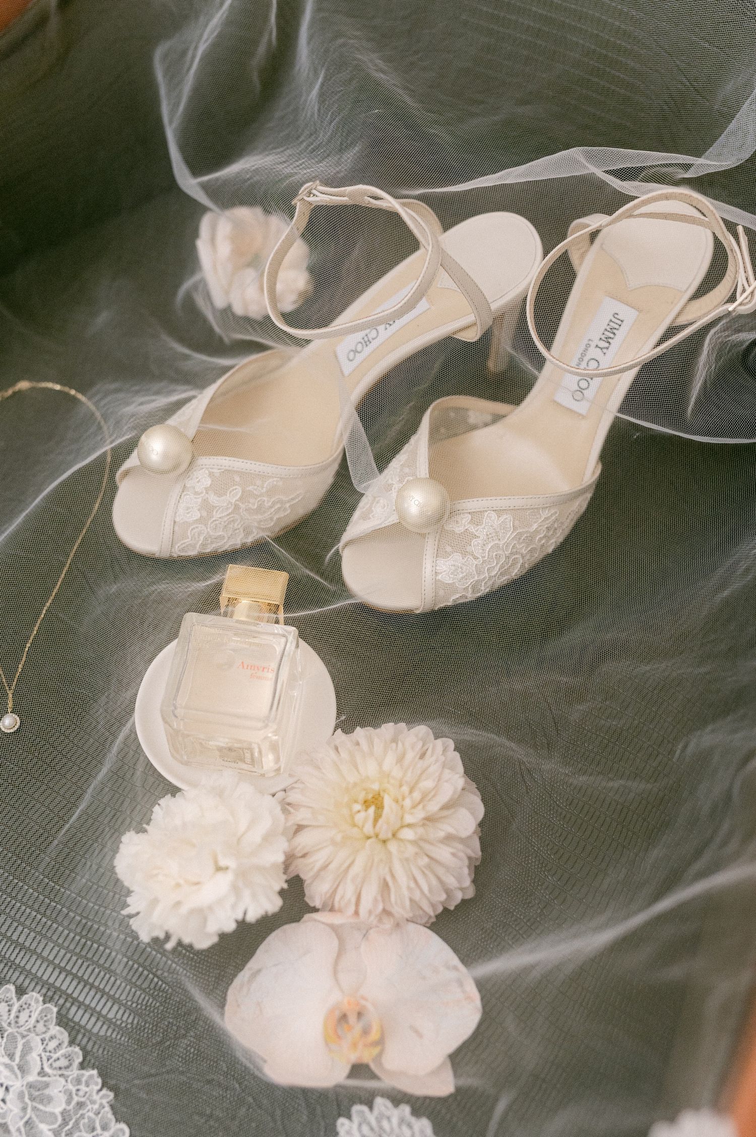 Elegant white bridal shoes with lace details displayed alongside a perfume bottle and delicate white flowers on tulle fabric.