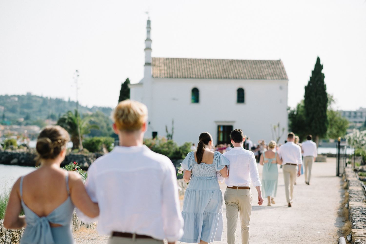 Wedding guests walking along a path towards a white Mediterranean church with cypress trees.
