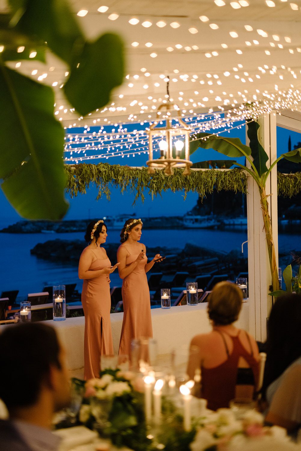Sunset wedding reception with string lights and ocean views as speakers address the guests.