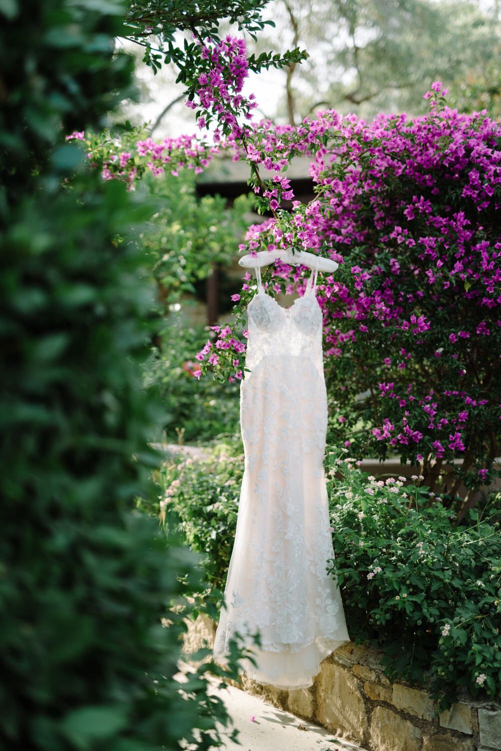 White wedding dress hanging against vibrant purple bougainvillea flowers in garden setting.