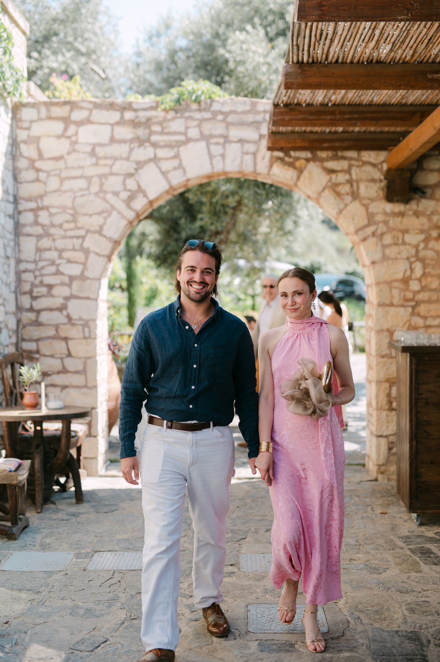 A couple walks together under a rustic stone archway covered patio space set among peaceful greenery.