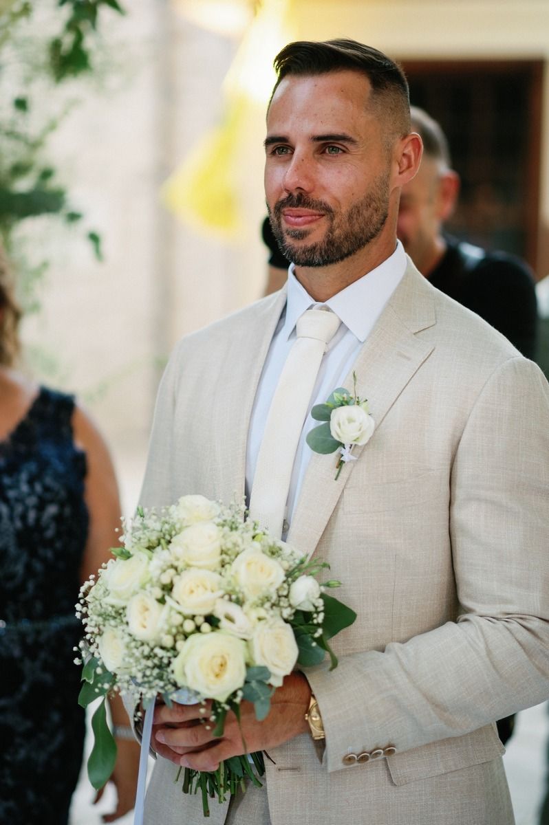 Wedding attendant holding white rose bouquet wearing light beige suit.