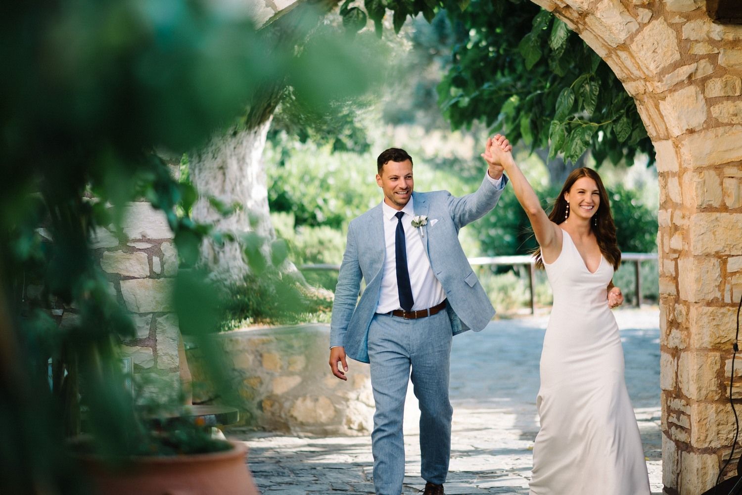 Newlyweds walking hand in hand under a stone archway surrounded by lush garden scenery.
