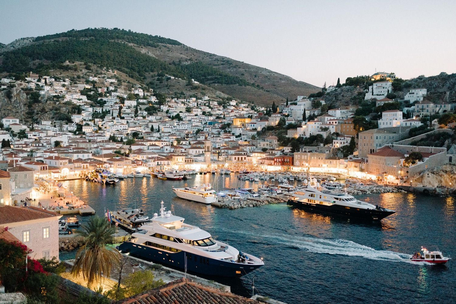 Aerial view of Mediterranean coastal town with boats docked in illuminated harbor at dusk.