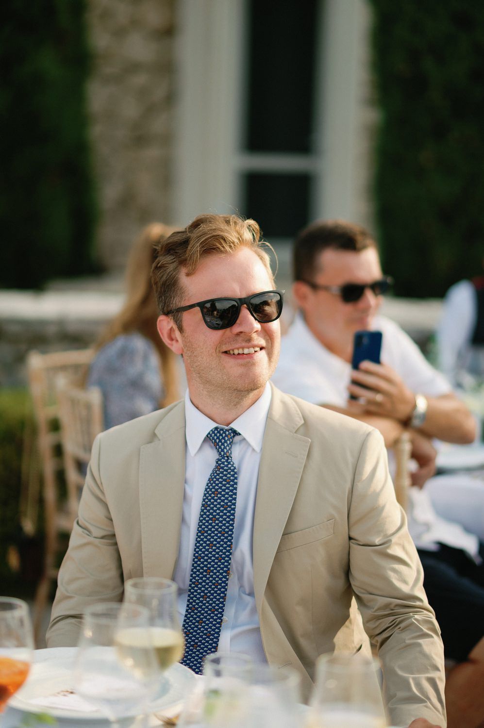 Guest in beige suit and sunglasses smiling at outdoor reception.