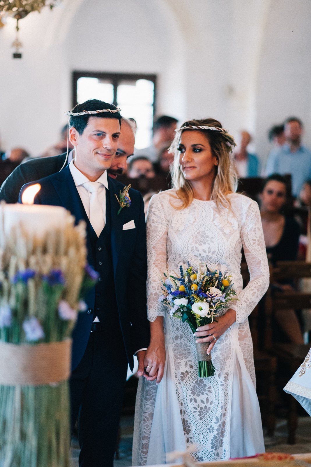 Bride in long-sleeved lace gown and groom in navy suit during Greek church ceremony.