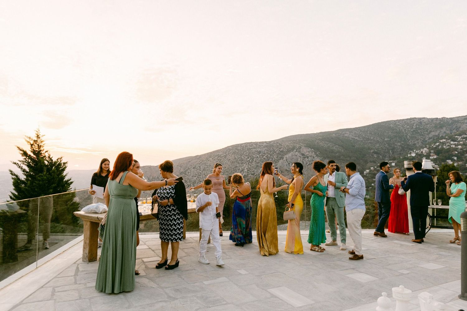 A group of people in formal attire gather on a mountaintop terrace for an outdoor event at sunset.