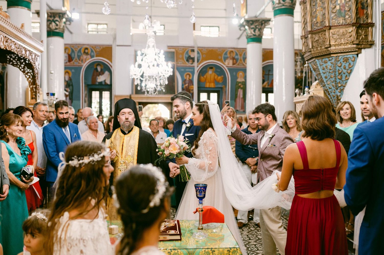 Elegant wedding ceremony in an ornate venue with crystal chandeliers and decorative columns surrounded by guests.
