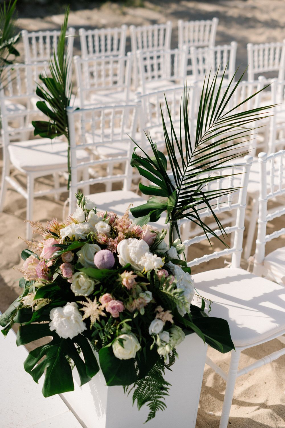 White chiavari chairs decorated with tropical flowers and palm leaves for beach wedding.