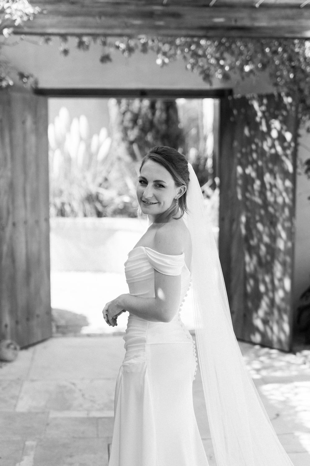 A monochrome portrait of a bride smiling under a wooden archway with natural light streaming through.