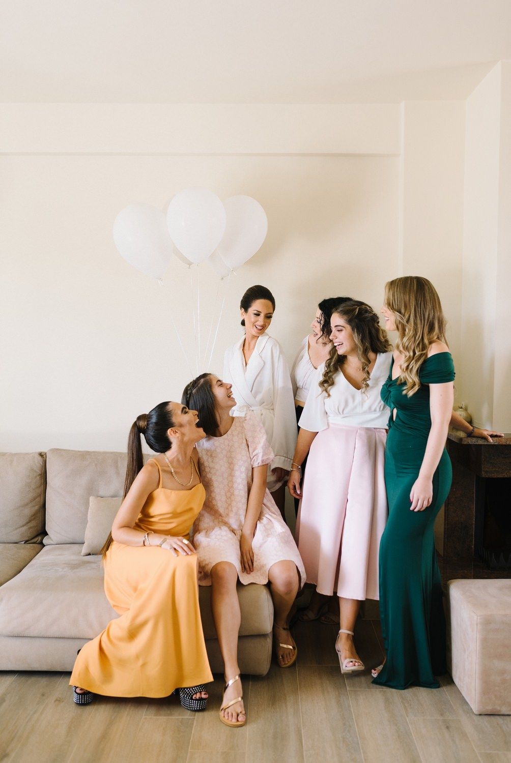 Bridal party in elegant dresses gathering on couch with white balloons in background.