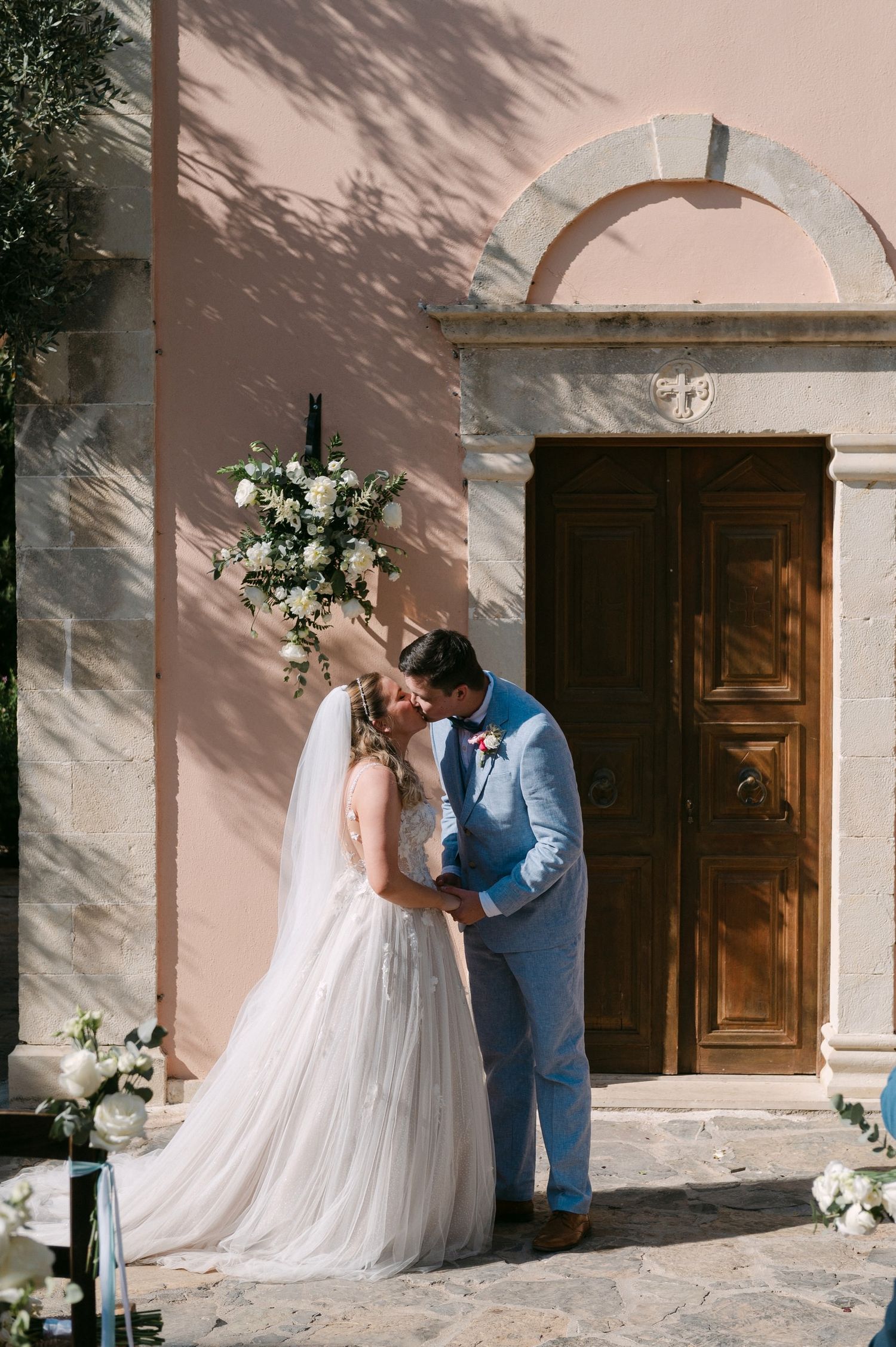 Romantic wedding couple shares tender moments near rustic wooden doors at a blush-colored Mediterranean style venue.