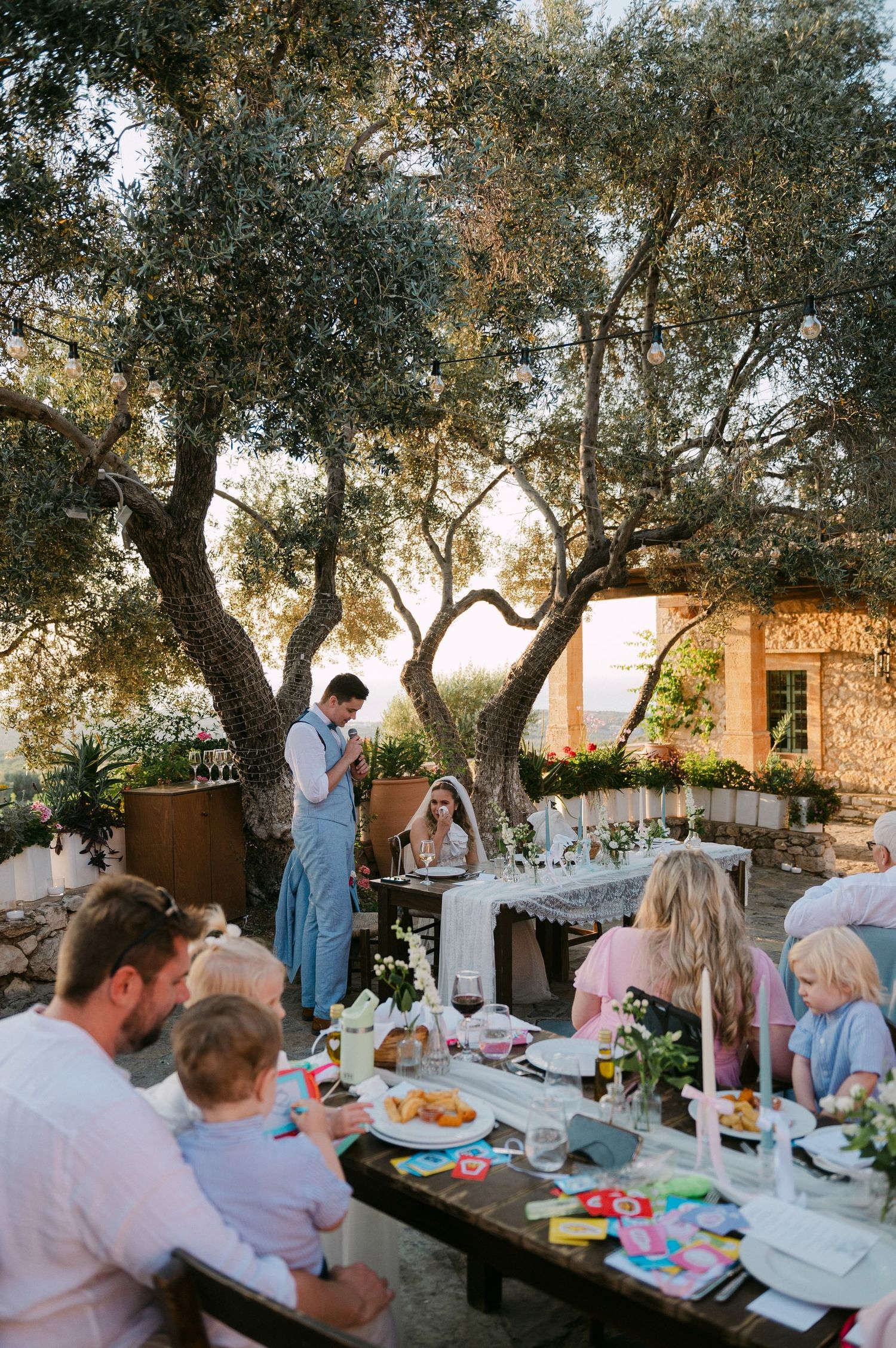 Casual outdoor dinner party under olive trees at sunset with table settings and glowing string lights above.