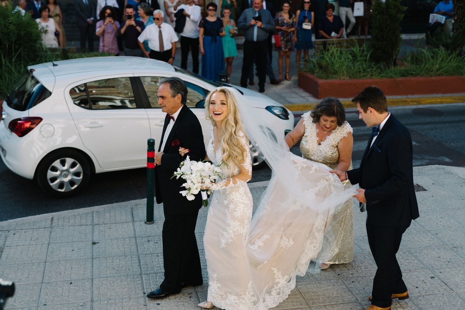 Wedding party helping bride with flowing dress as they walk outside by a parked car.
