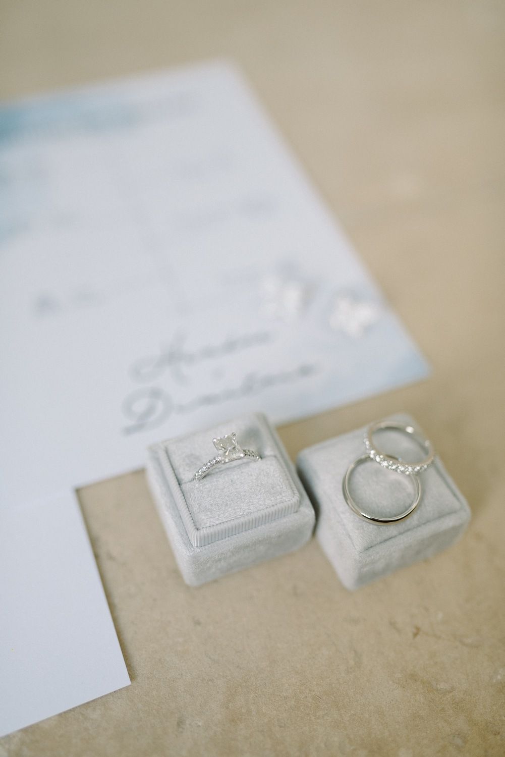 Wedding rings displayed in gray velvet boxes next to an invitation on a light surface.