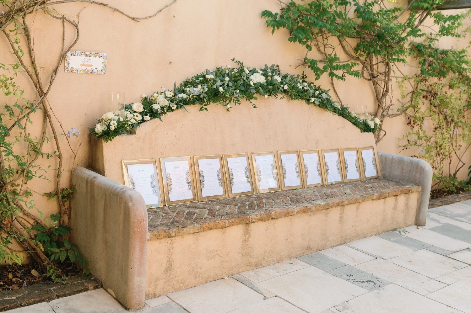 Stone bench with wedding seating cards arranged in a row under a floral arch.