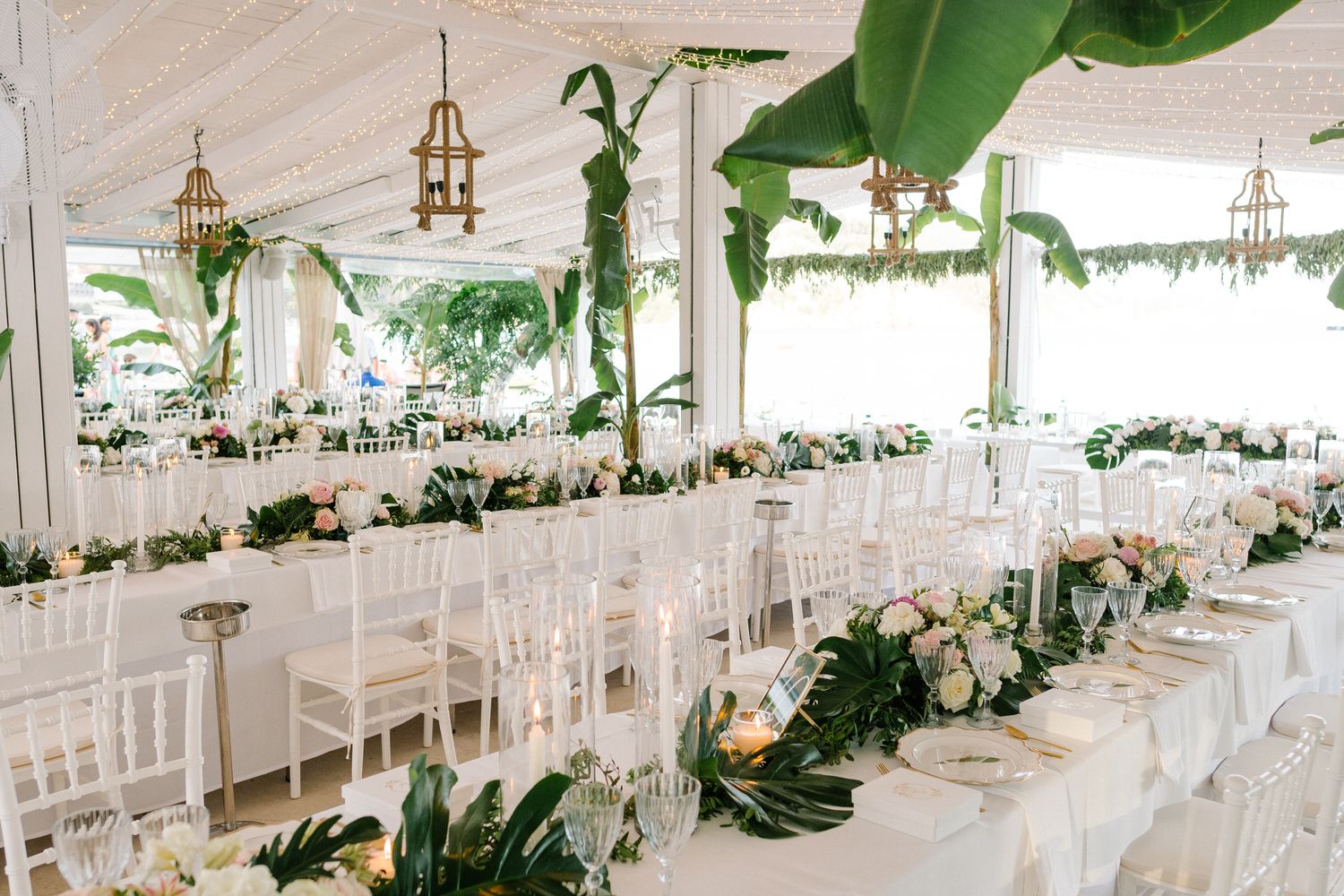 Elegant white wedding reception hall with tropical greenery, string lights and white chairs.