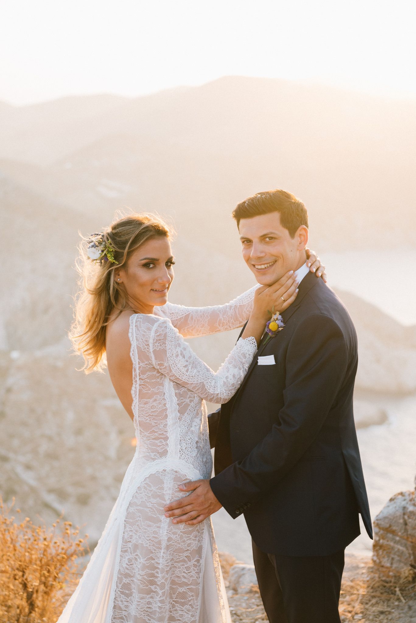 A couple in wedding attire poses together on a mountaintop at sunset with golden light and scenic views.