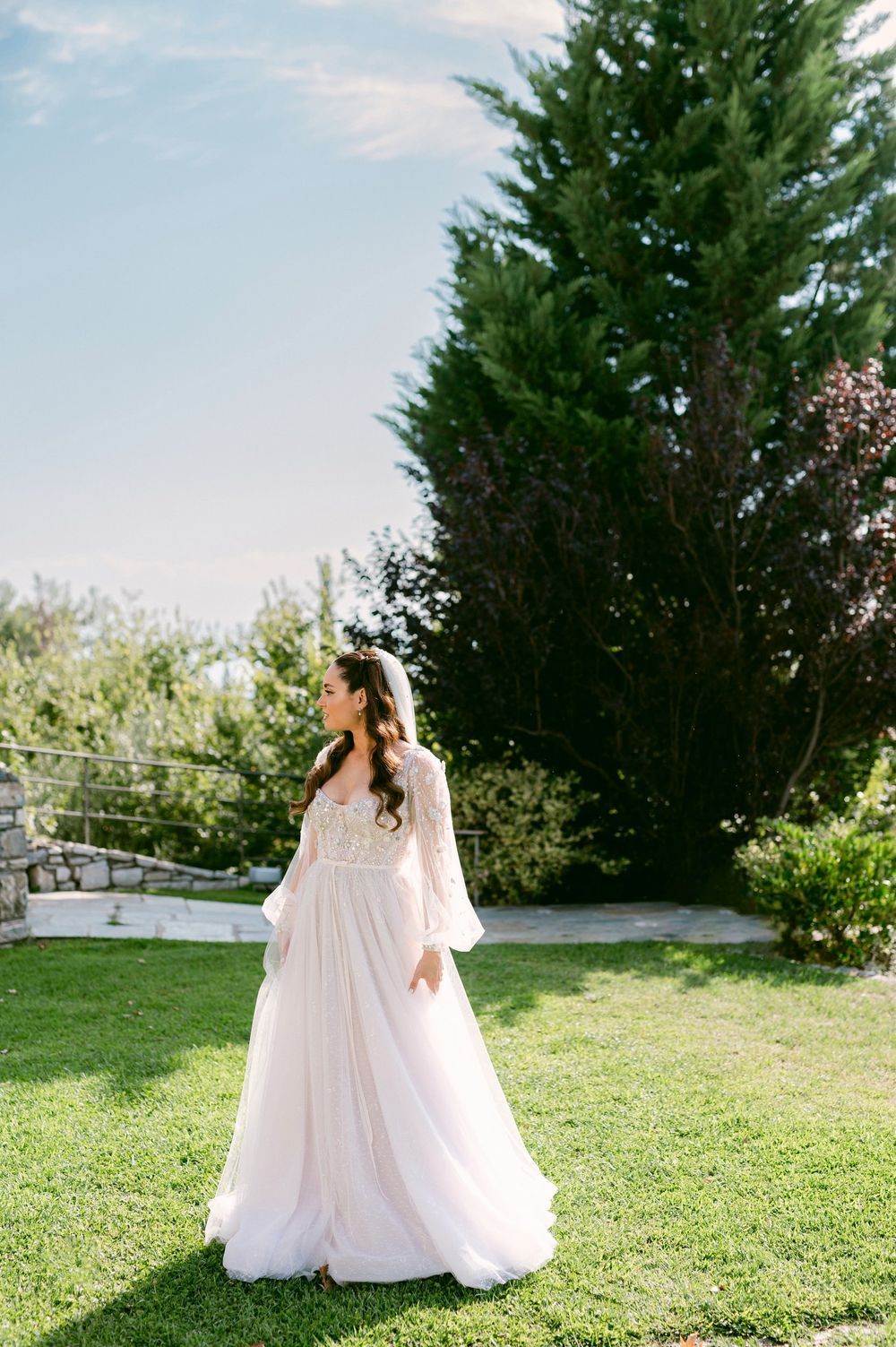 A person in a flowing white dress walks gracefully across a sunlit grassy hill with evergreen trees in the background.