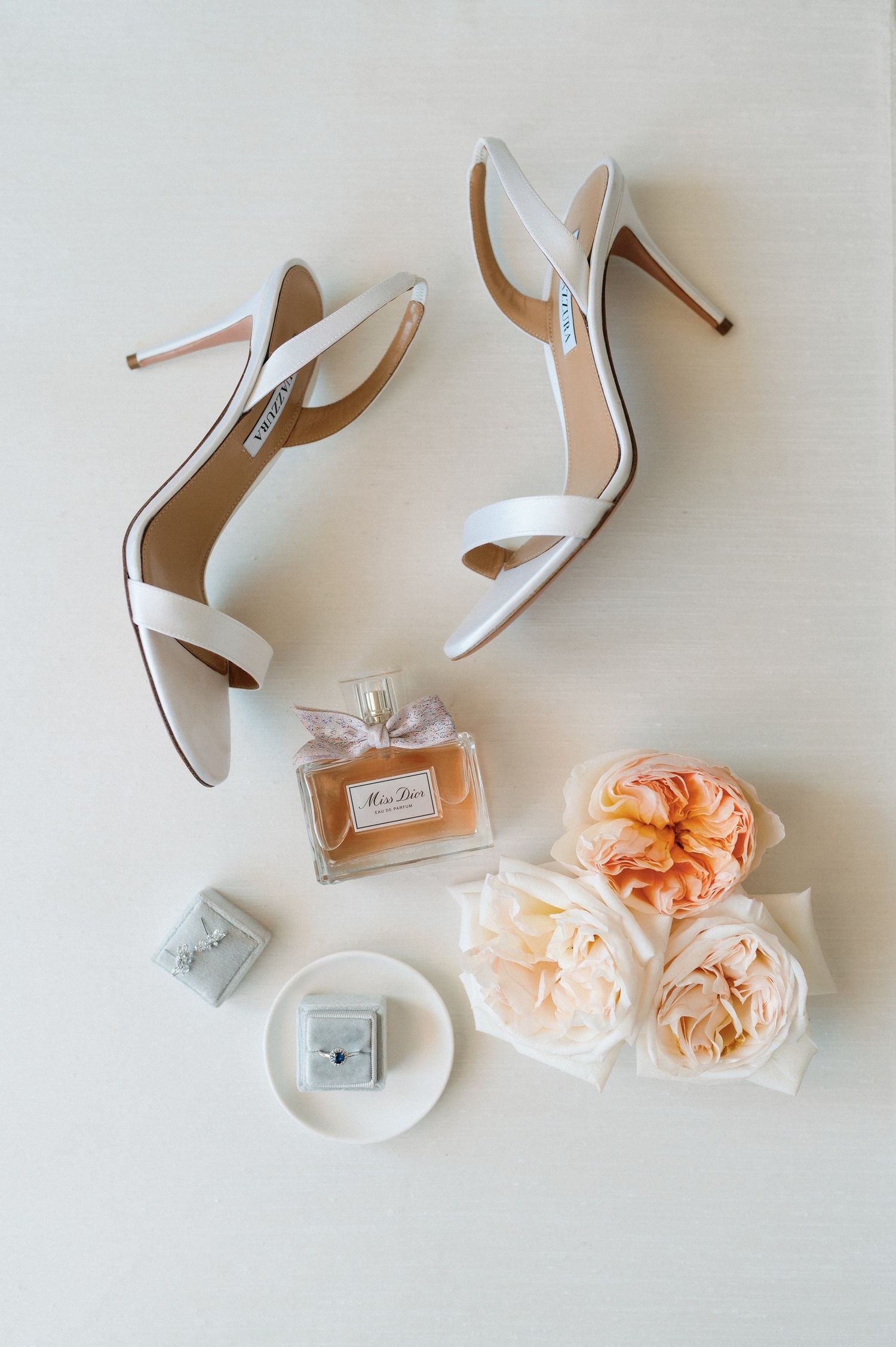 Elegant white bridal shoes arranged with peach garden roses and perfume bottle on light background.