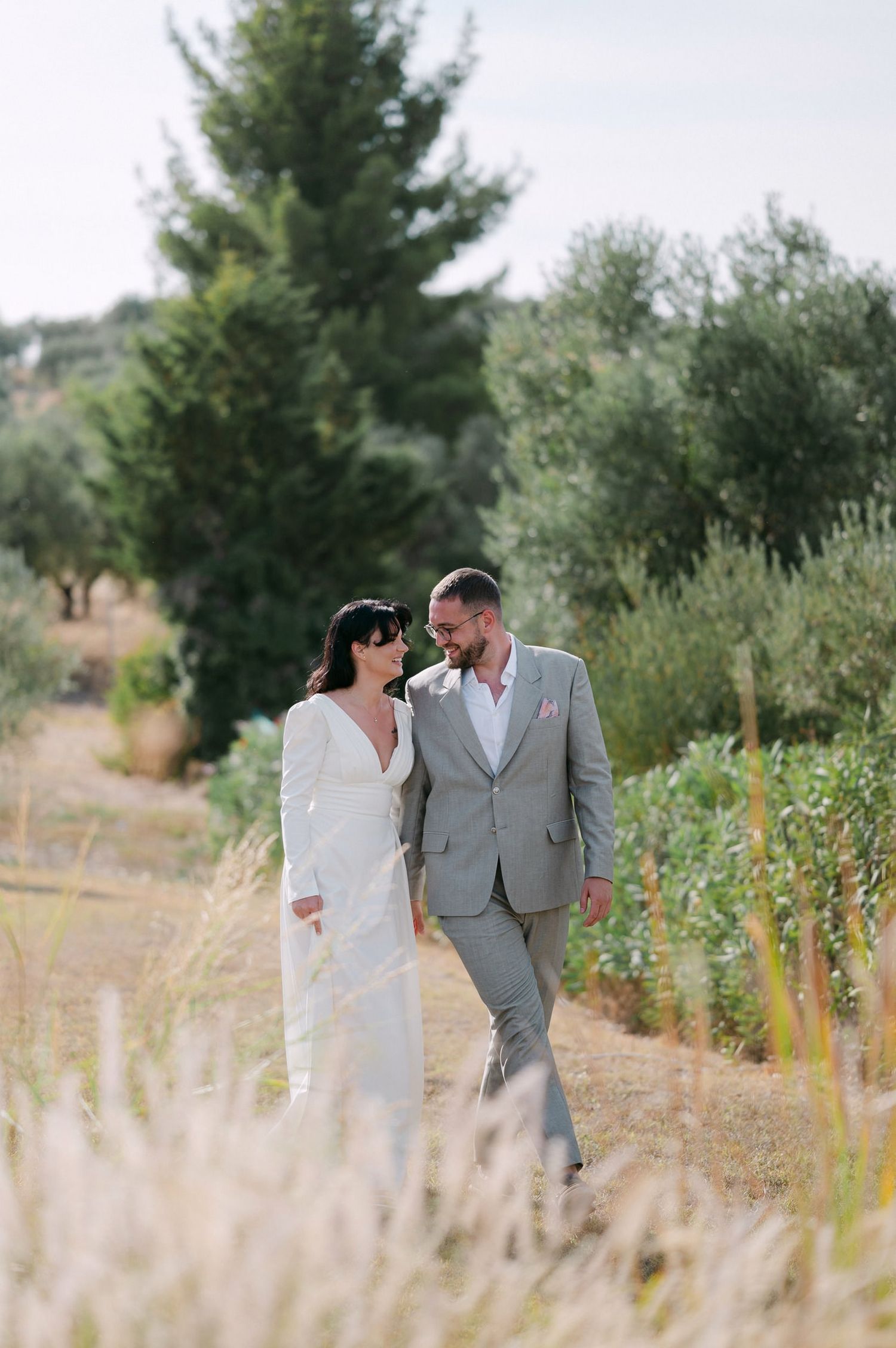 A couple in wedding attire walks together through a field of tall grass with evergreen trees in the background.