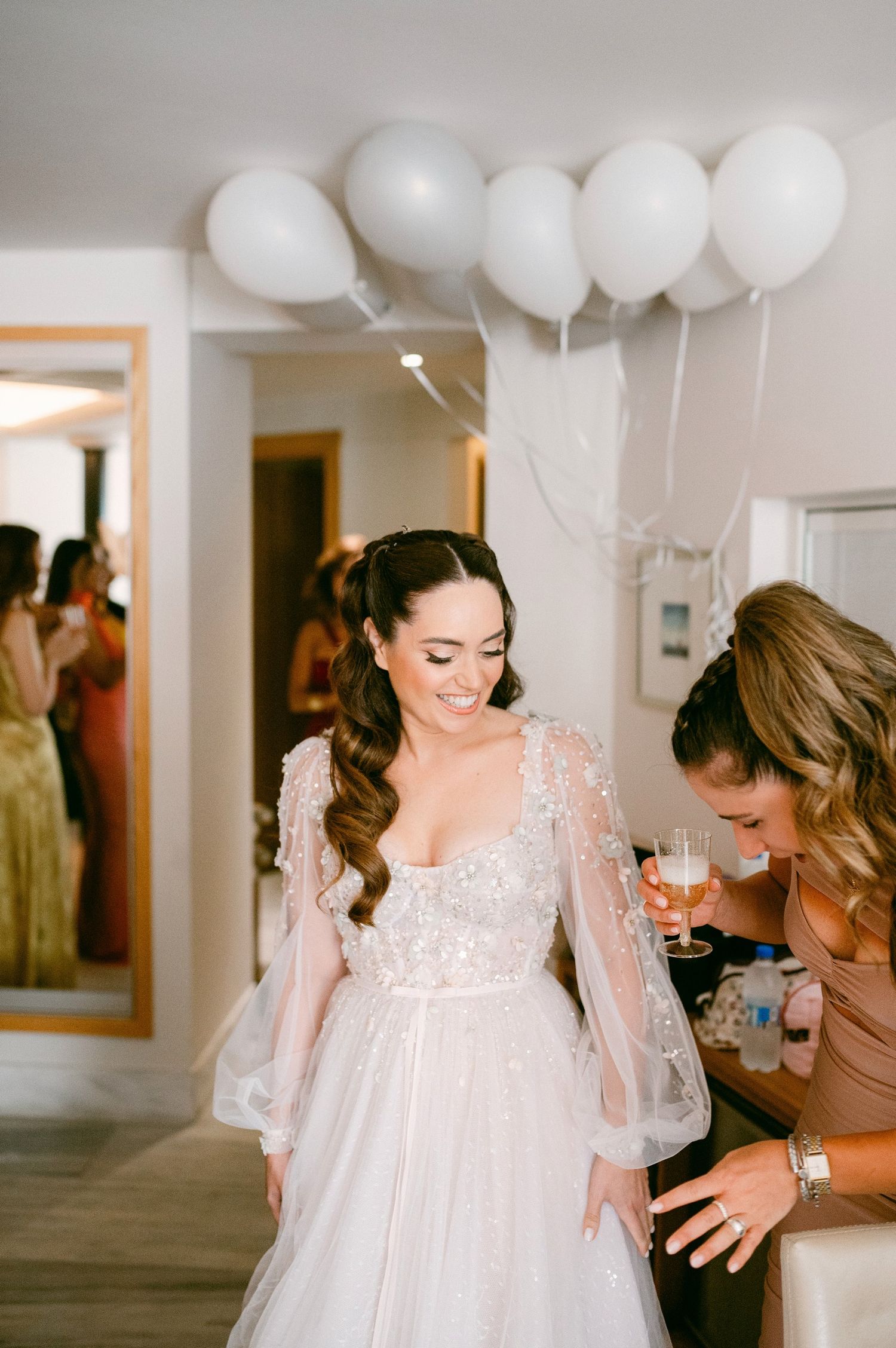 A bride in a flowy lace wedding dress with billowing sleeves stands in a room decorated with white balloons.